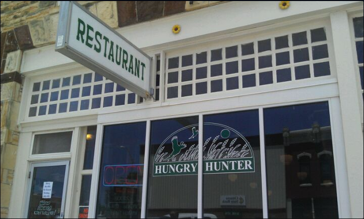 color photo of the front of the Hungry Hunter Restaurant in Lincoln, Kansas. The photo shows the large front windown with a painted sign of birds flying up through stalks of praire grass. The red open sign is on. The front window shows the reflection of three story buildings across the street.