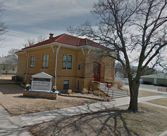 color photo of the Lincoln, Kansas, Carnegie Library. A limestone building with a red tile roof and red double front doors. Cement steps leading up to the doors. A new addition is visible on the back.