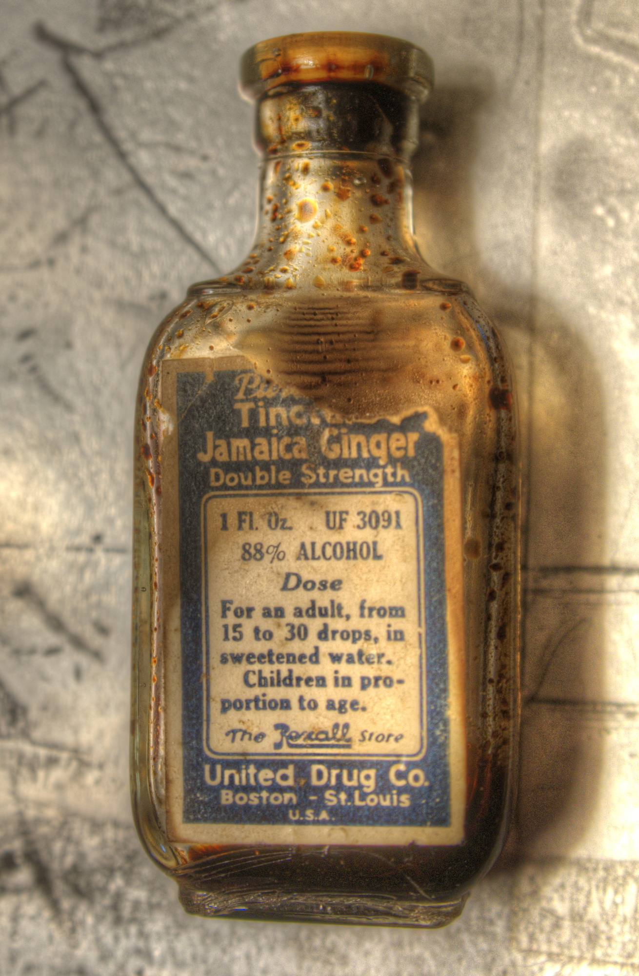 very old glass bottle with cork stopper. Front label is blue and whiteand a small portion of the upper right hand corner of the label is torn away. But the labeling is still there and reads, "Jamaica Ginger Double STrength. 1 Fluid oune, 88% alcohol. Dose for an adult, from 15 to 30 drops, in sweetened water. Children in proportion to age. The Rexall Store. United Drug Co.l Boston - St. Louis U. S. A. The glass is clear and dark reddish-brown droplets and residue are visible inside the bottle.and reads